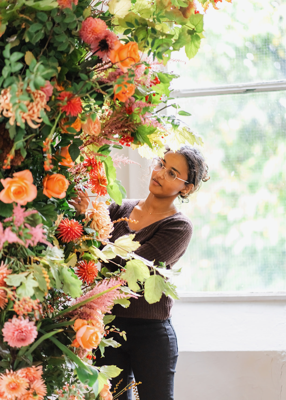 A woman with glasses arranging a colorful floral display indoors near a large window.
