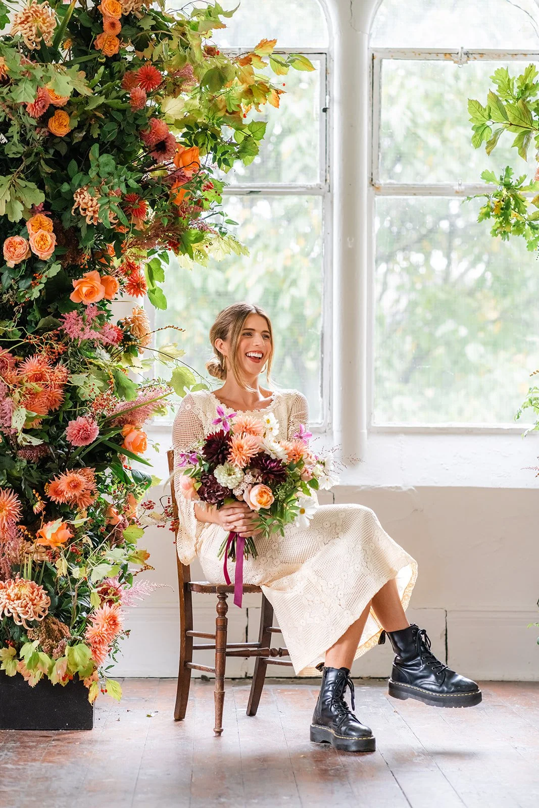 A woman sitting on a wooden chair holding a colorful bouquet of flowers, smiling, with a large window and a lush green landscape outside, and a floral arrangement on her left.