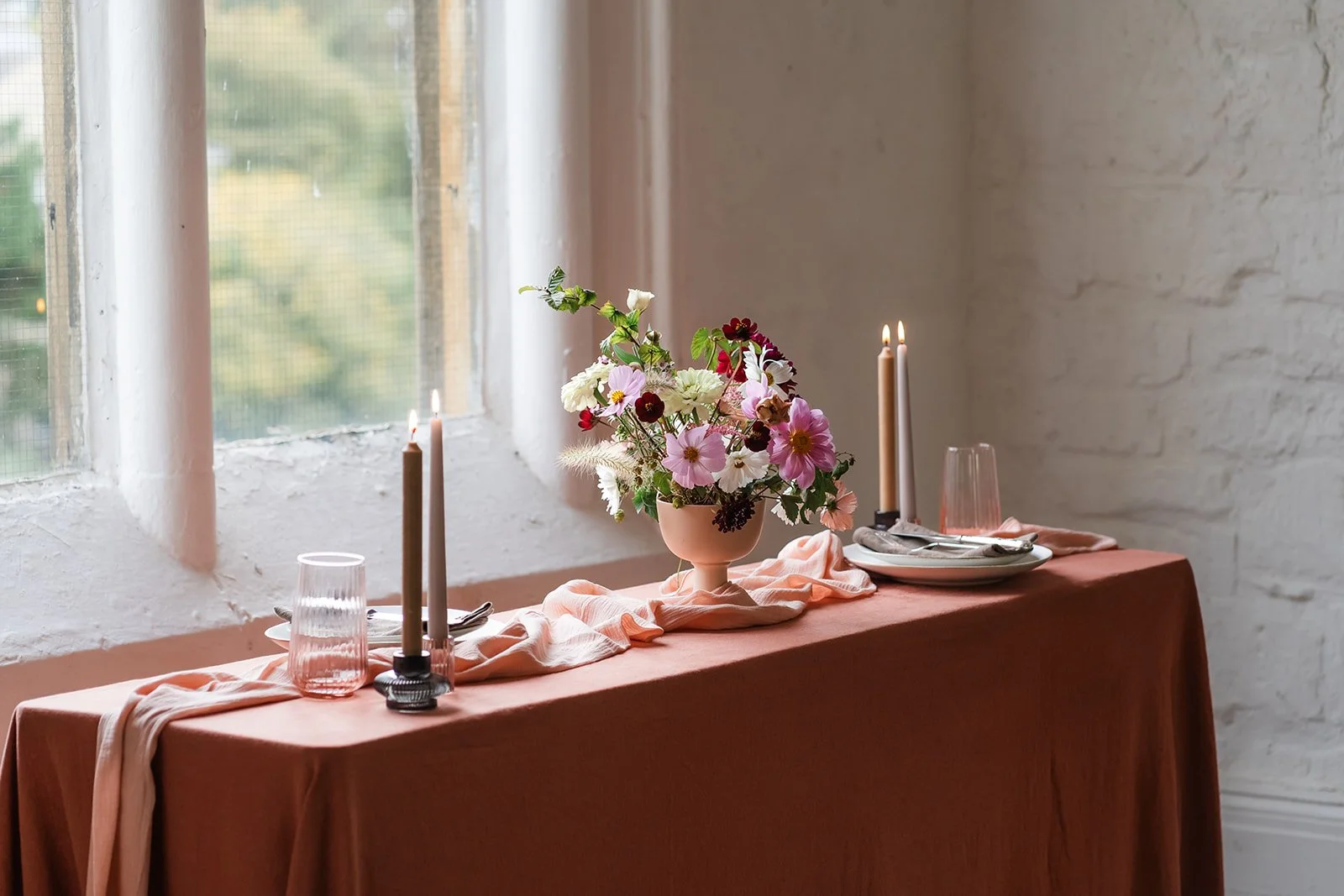 A table set for a meal with a pink tablecloth, a floral centerpiece with pink, white, and dark red flowers, two tall candles, glasses, and plates, near a window with natural light.