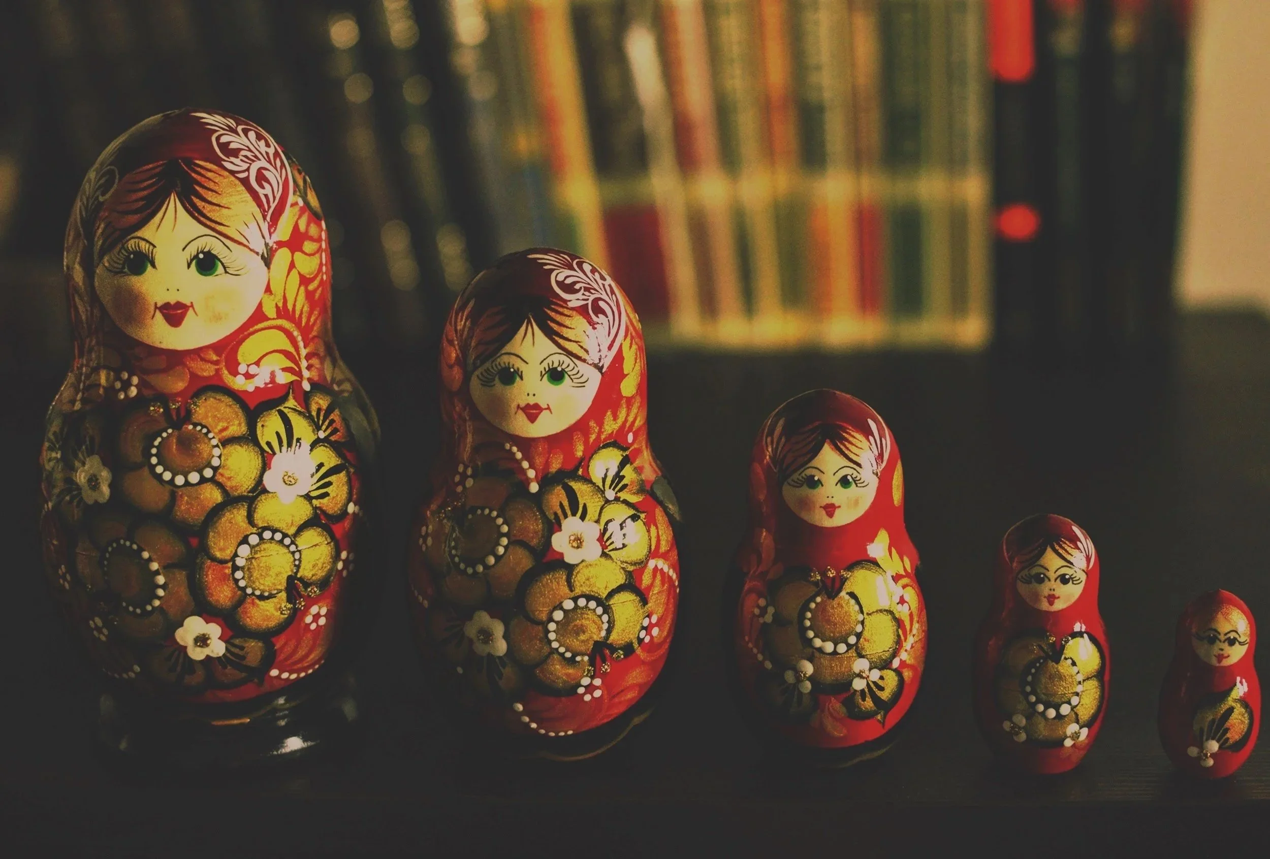 A set of five traditional Russian nesting dolls, painted with red, gold, and black floral designs, arranged in decreasing size from left to right, with a blurred bookshelf in the background.