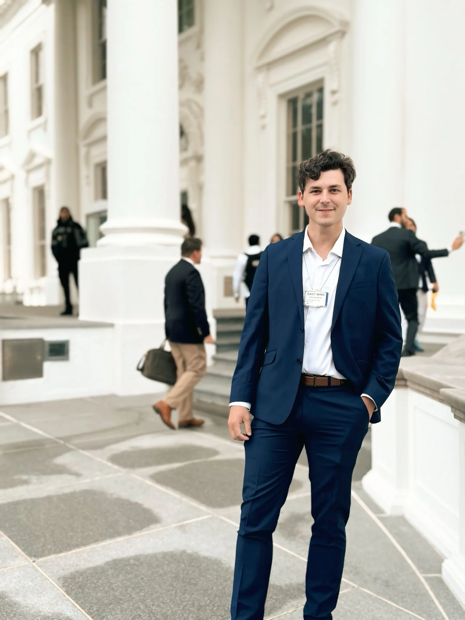 Cam Higby standing in a navy blue suit and white shirt standing outside of the White House. Several people are in the background, some walking, some talking, some taking photos.