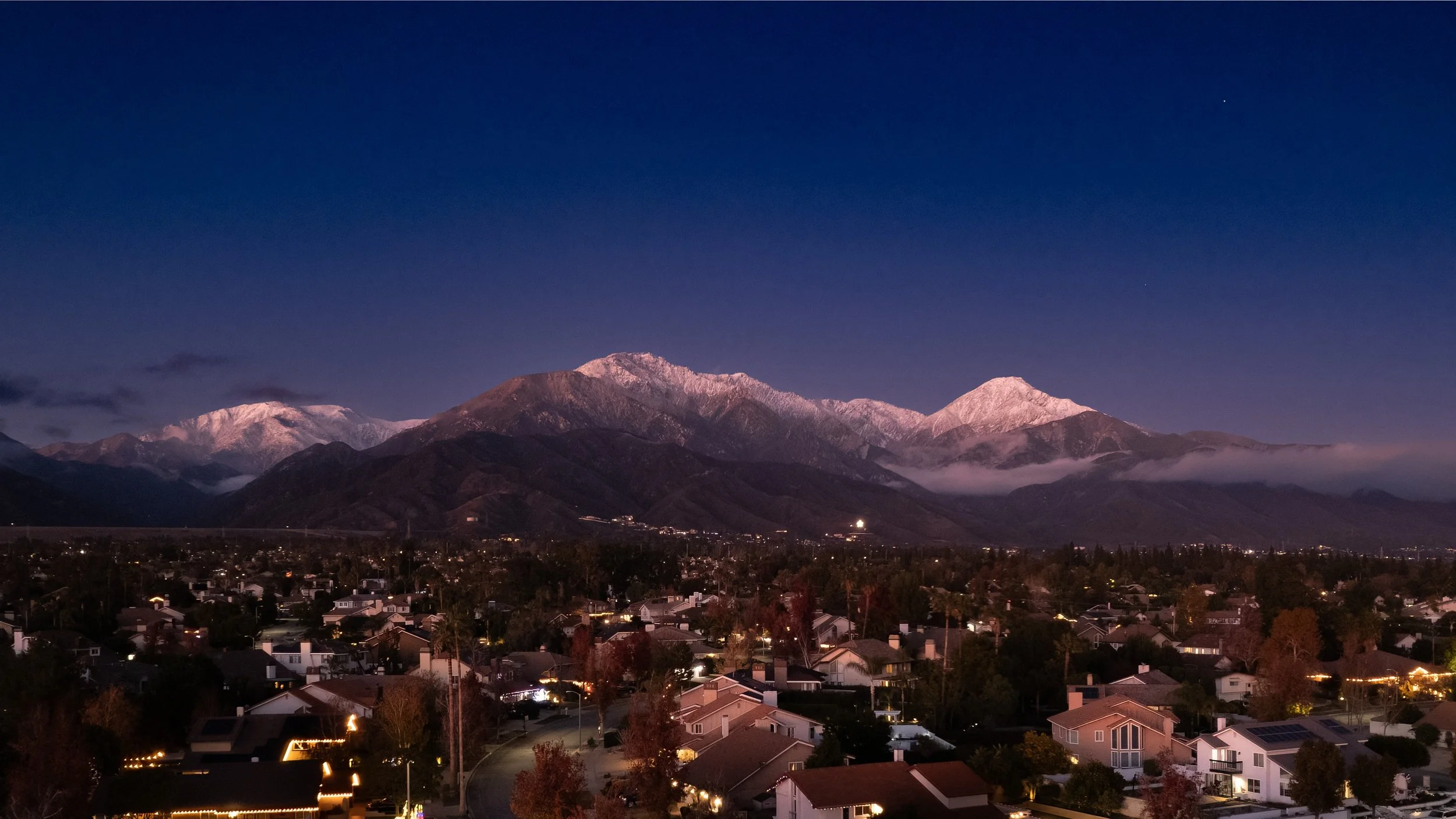 Dramatic twilight drone view of the Upland cityscape with a snow-capped Mt. Baldy (Mount San Antonio) in the background. High-end aerial real estate photography in North Upland and San Antonio Heights by SoCal Home Photo.