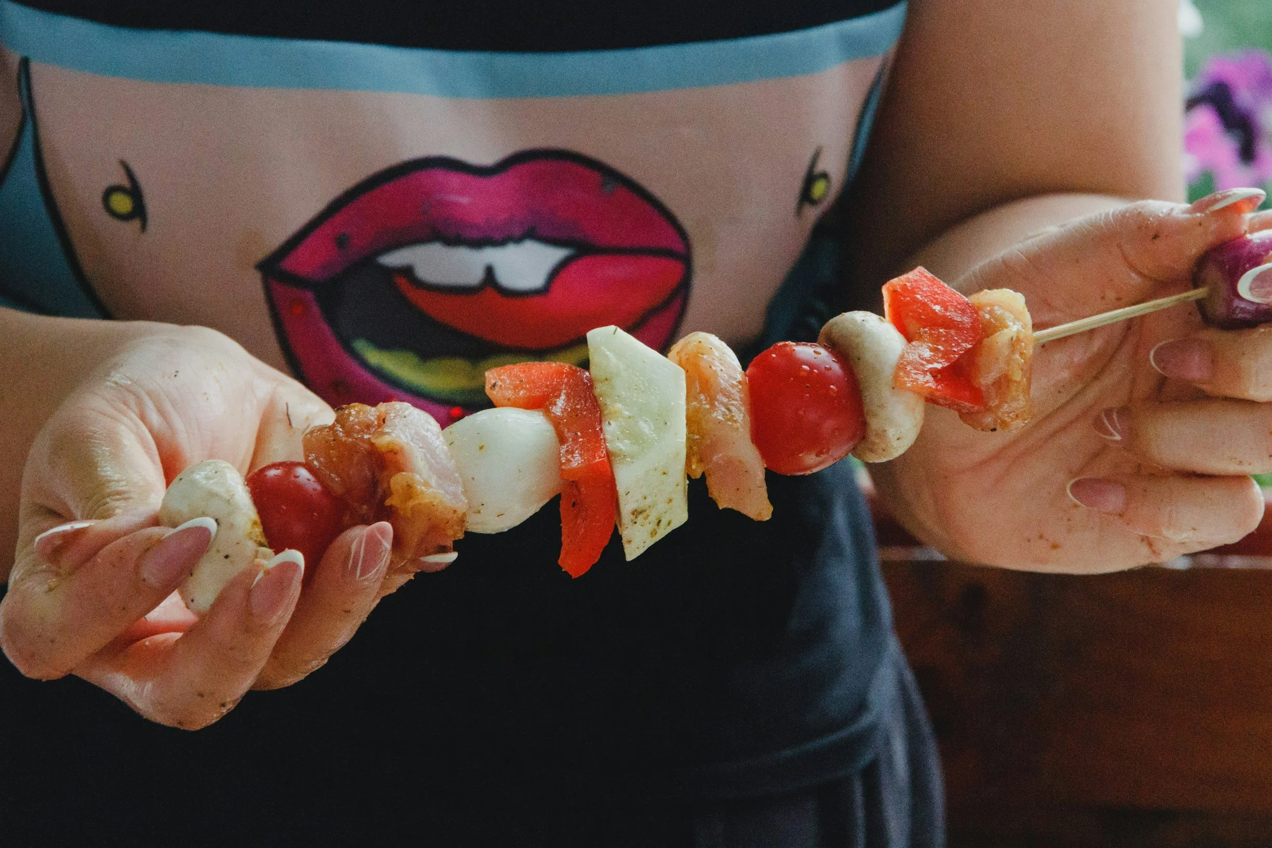 Person holding a skewer with cherry tomatoes, mozzarella, and red bell peppers, wearing a shirt with a comic-style mouth graphic.