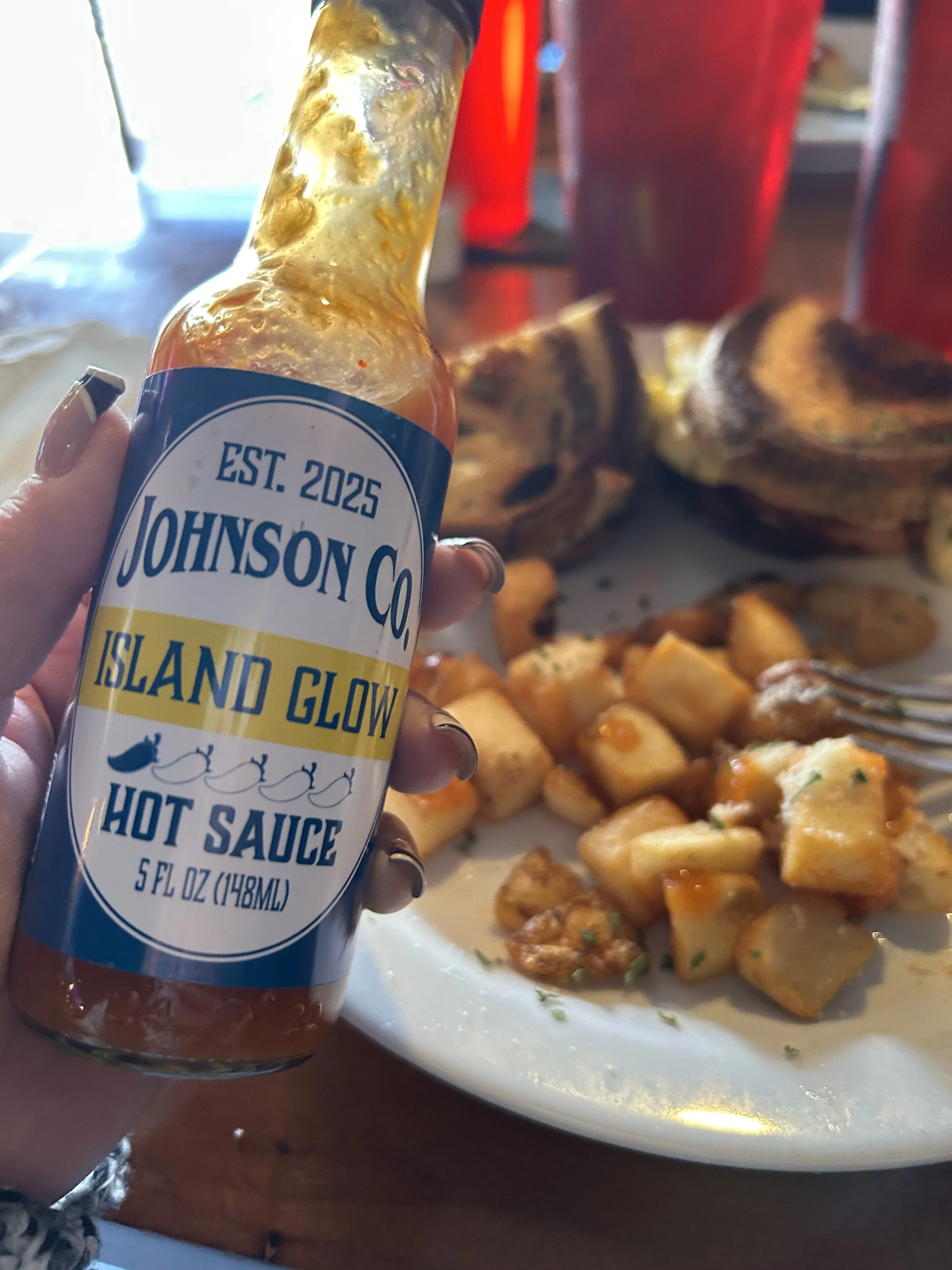 Bottle of Johnson Co. Island Glow hot sauce held by a person's hand, with a plate of roasted chicken and fried potatoes in the background.