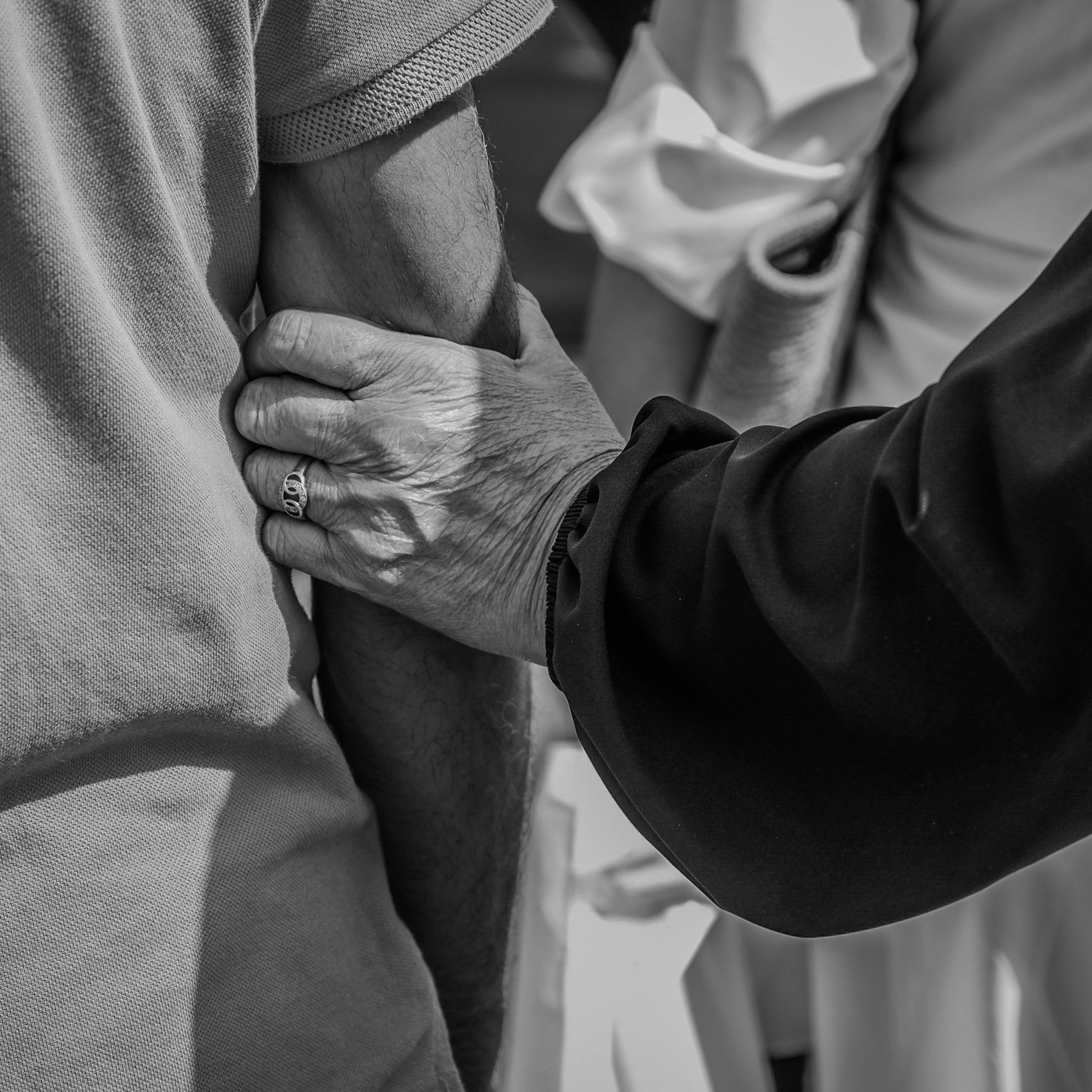 An elderly person's hand holding another person's arm during a medical vaccination or blood draw, with a close-up focus on the hand and arm in black and white.