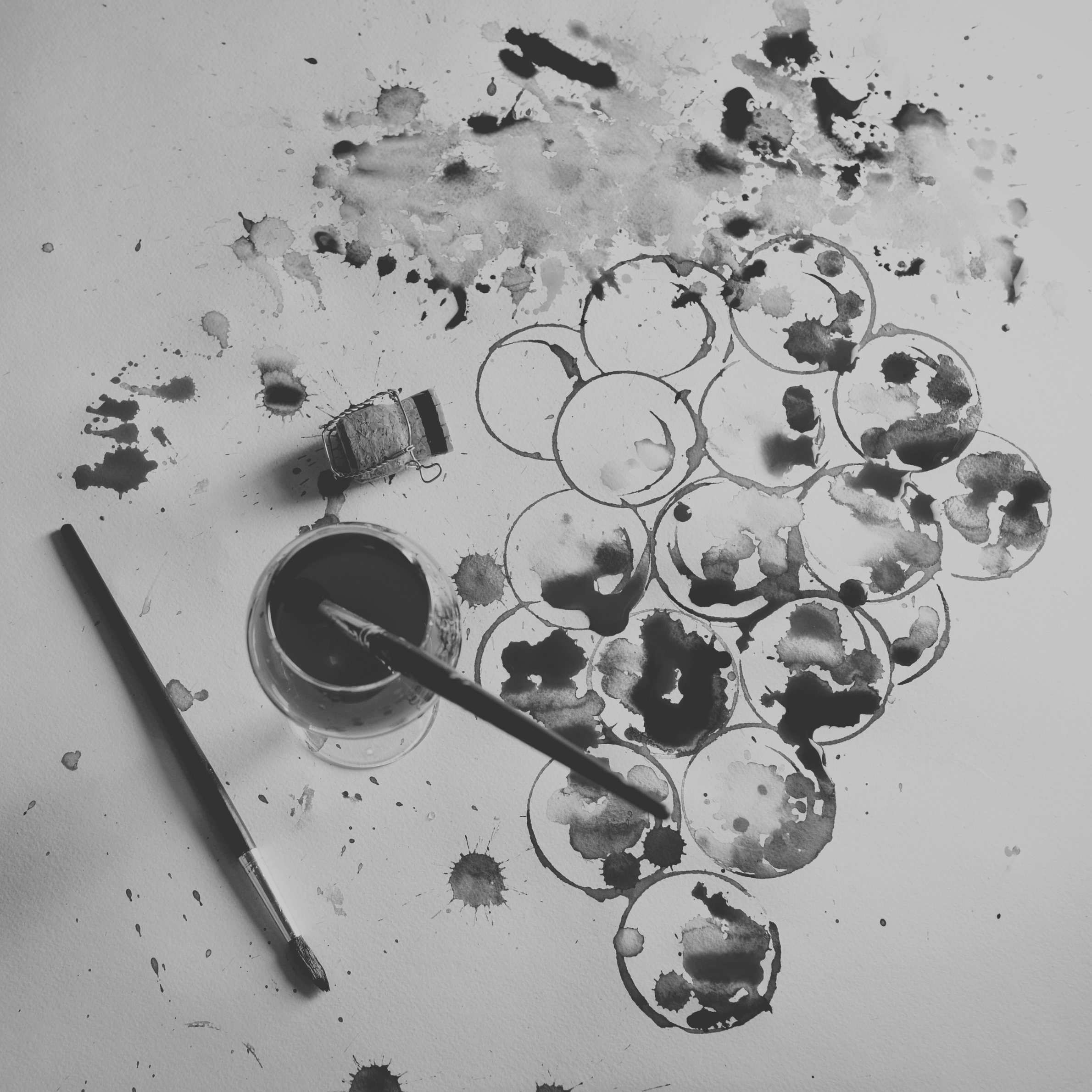 Art supplies on a table including an open ink bottle with a brush dipped in, a paintbrush, and a sheet of paper with watercolor circles and splatters.