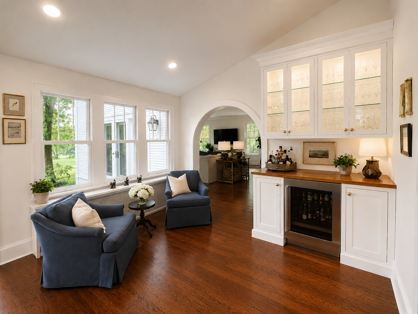A cozy living room with two blue armchairs, a small black side table, and a wooden fireplace with a white cabinet above it. The room has large windows, a patterned red rug, and decorative items on the shelves and walls.
