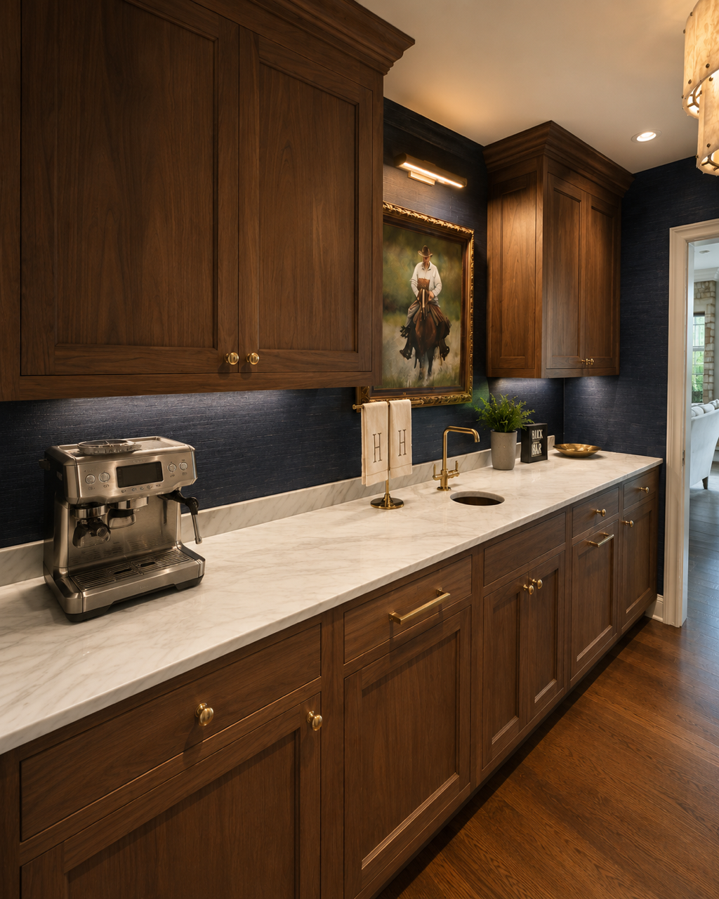 A kitchen with wooden cabinets, a marble countertop, and a framed painting of a cowboy on a horse hanging on a dark wall. There are bottles and bar utensils on the left side and books and a small sculpture on the right side of the countertop.
