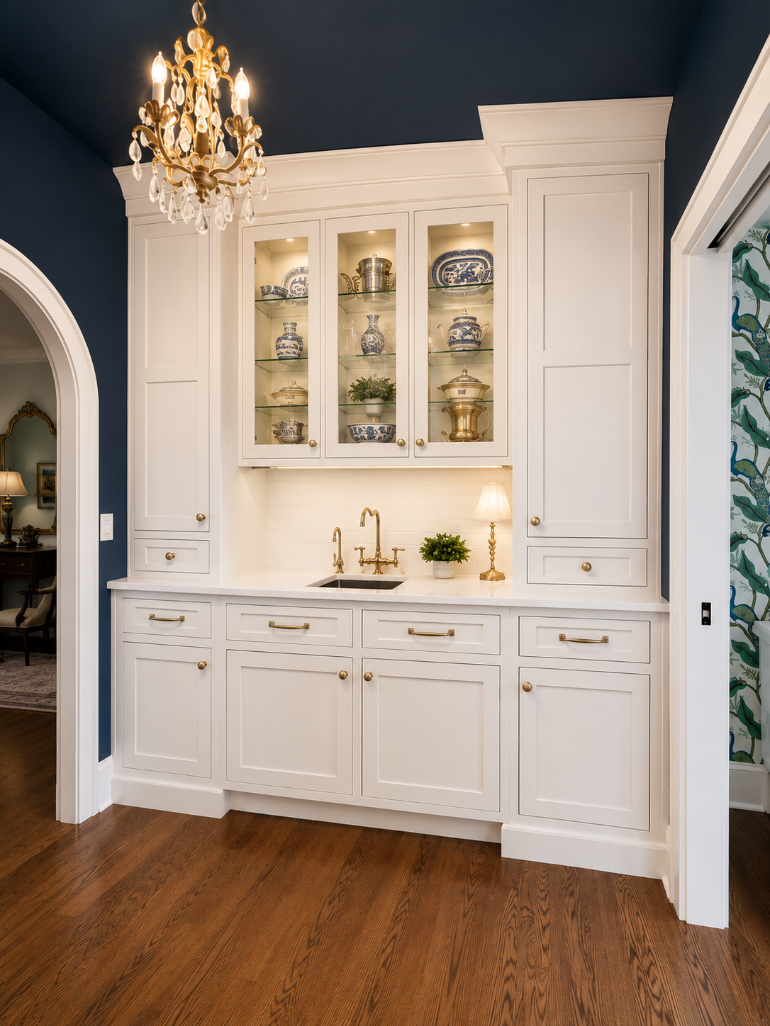Kitchen with white cabinetry, glass-front upper cabinets displaying dishes and decor, a small sink, a coffee maker, and a table lamp. Decor includes a small plant and a pumpkin ornament.