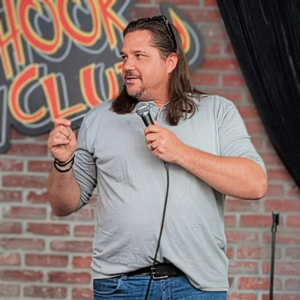 Man with long hair and beard performing stand-up comedy on stage, holding a microphone, with a brick wall and colorful sign in the background.