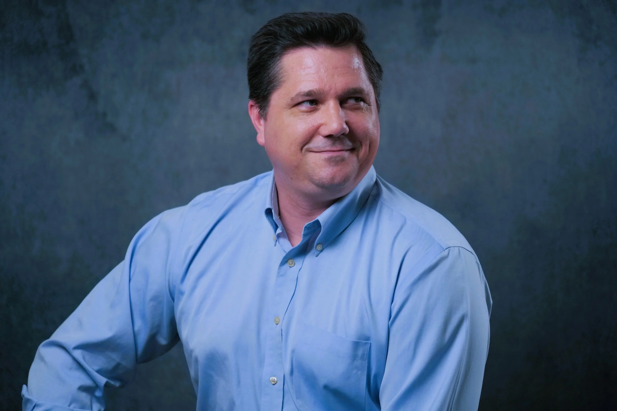 A man in a light blue button-up shirt looking to his right with a slight smile, with a dark textured background.
