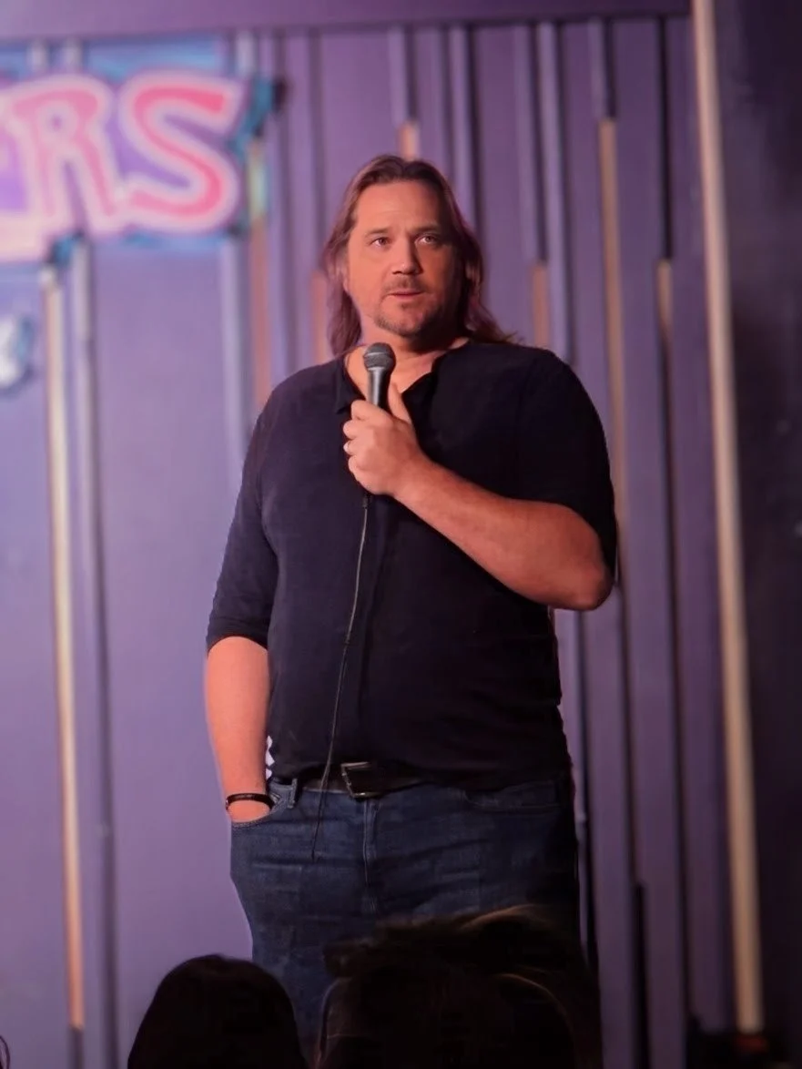 A man with long brown hair holding a microphone, standing on a stage in front of a purple backdrop with vertical lines, wearing a black shirt and jeans.