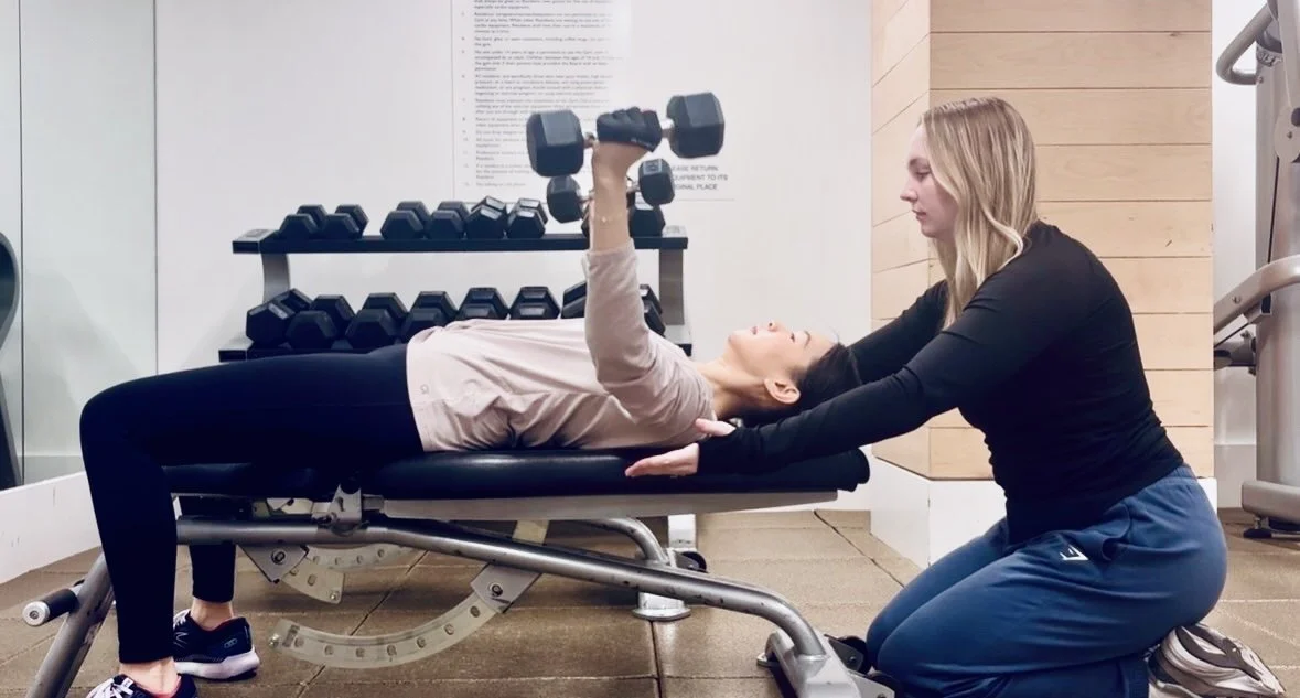 A woman in workout clothes performs a dumbbell chest press exercise on a bench while a trainer assists her with proper form at a gym.
