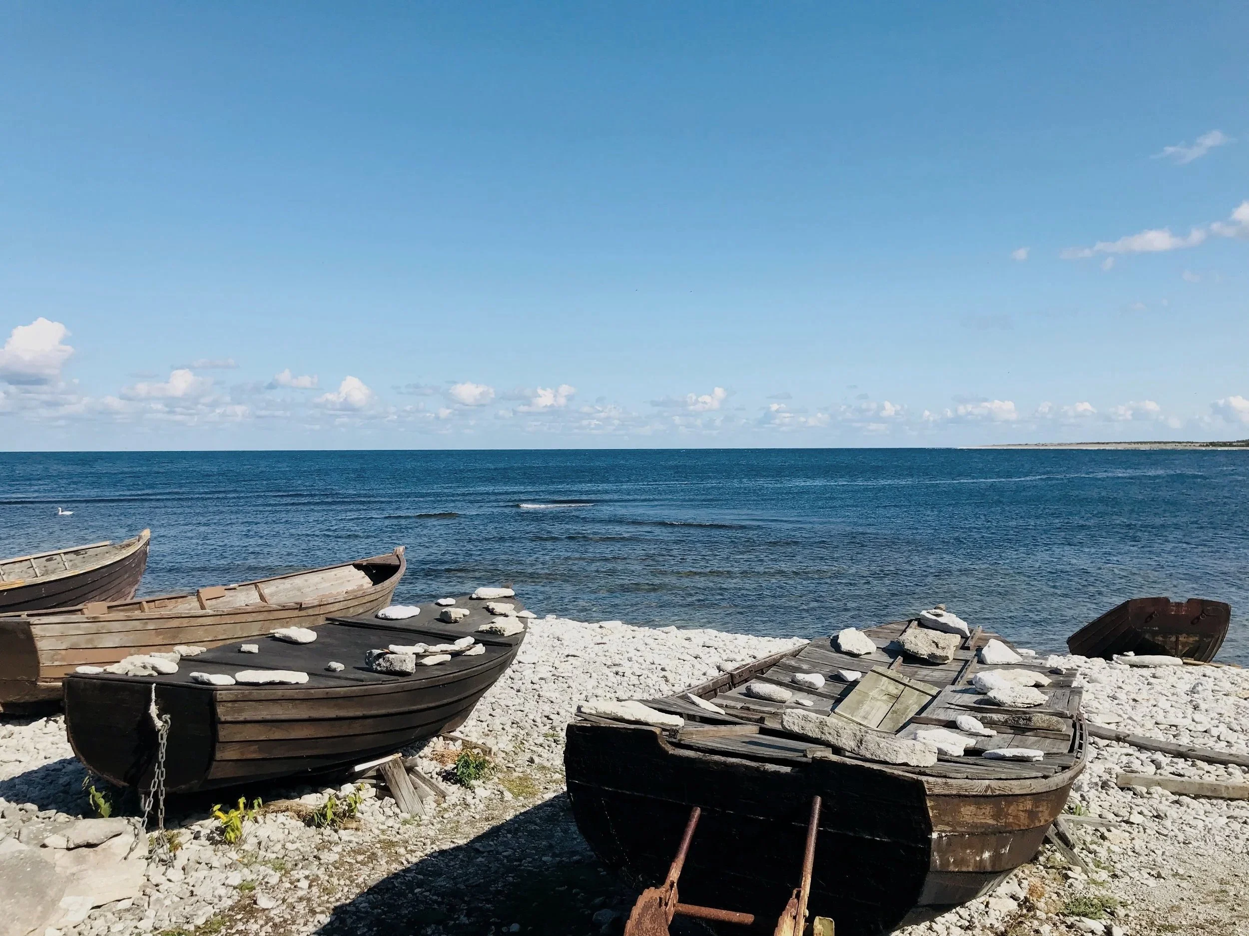 Vier alte, verlassene Holzboote liegen am Ufer eines Sees, mit Steinen bedeckt. Im Hintergrund ist das offene Wasser und ein blauer Himmel mit wenigen Wolken.