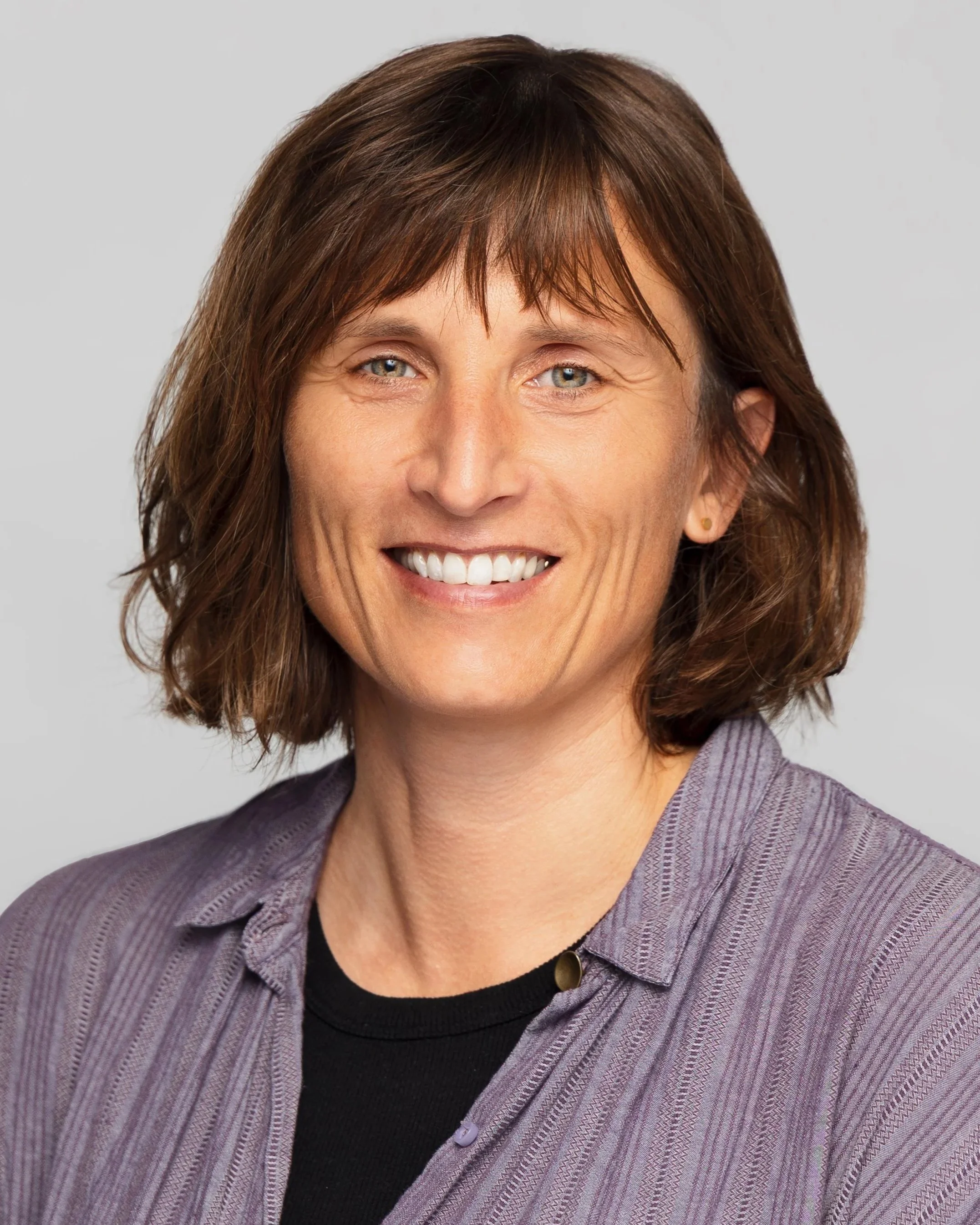 Porträt einer lachenden Frau mit braunen, lockigen Haaren, trägt eine lila Bluse und ein schwarzes Oberteil, vor hellem, neutralem Hintergrund.