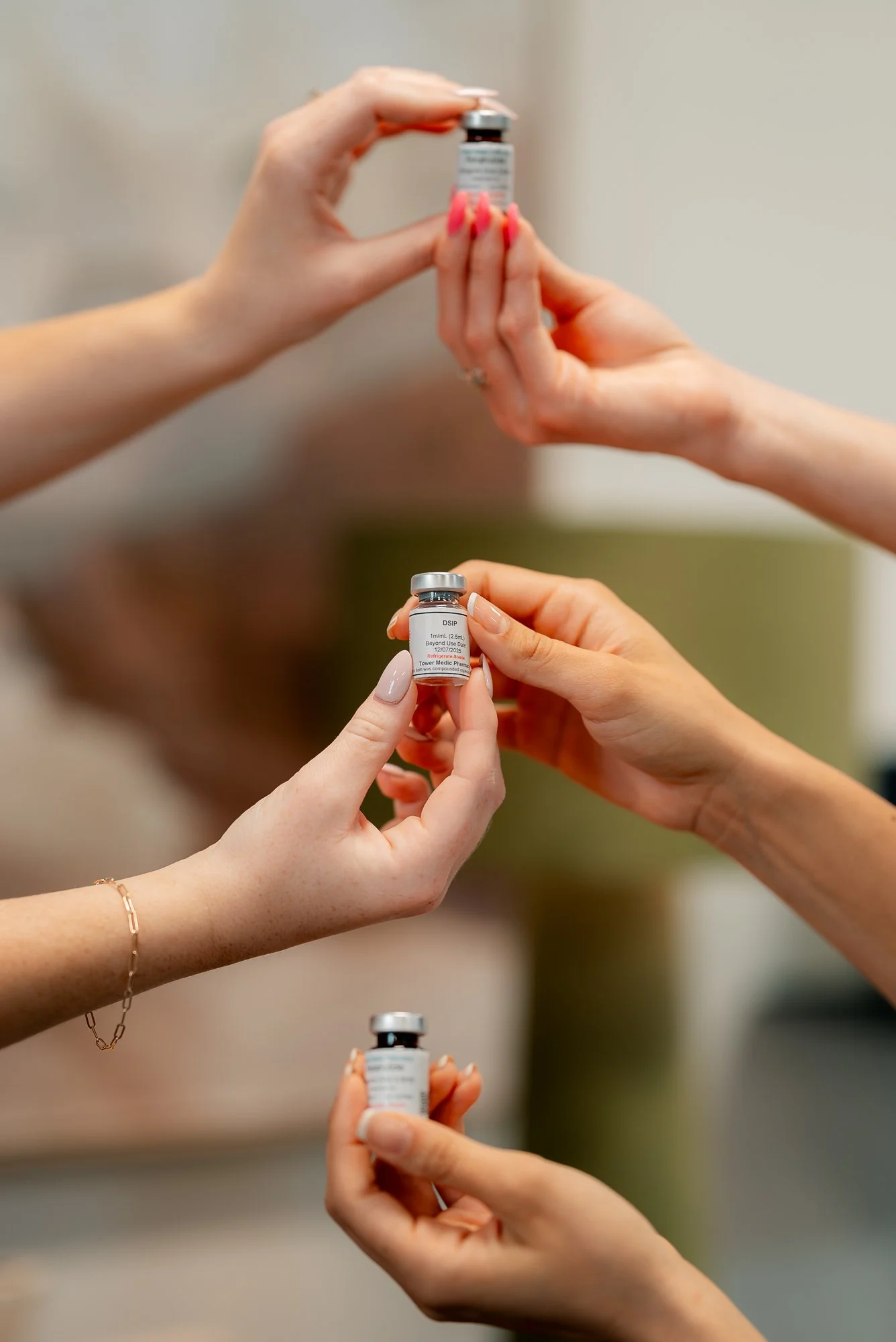 Three hands each holding a small vaccine vial, with two vials visible in the image.