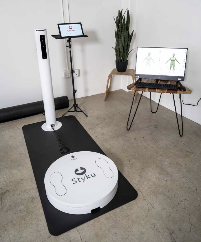 A medical or fitness scanning device with a large circular base labeled 'Styku' and footprints, a vertical stand with a monitor displaying the Styku logo, a computer with a monitor showing 3D body scans, a potted plant, and a black exercise mat on the floor.