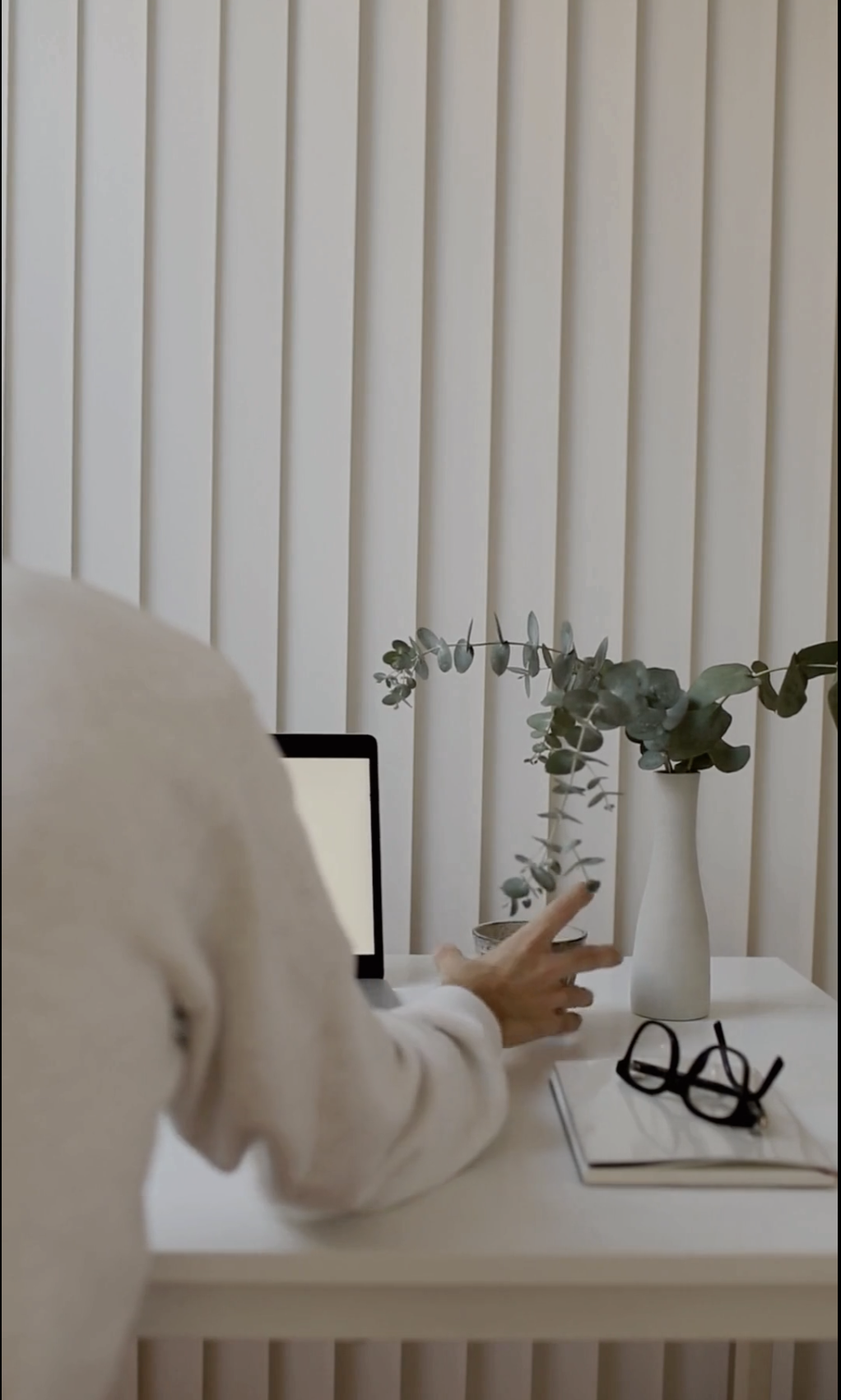 Person arranging plants in a white vase on a white desk with a laptop, glasses, and a notebook, with a white slatted wall in the background.