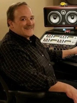 Man sitting at a music production desk with studio monitors and audio equipment, smiling at the camera.