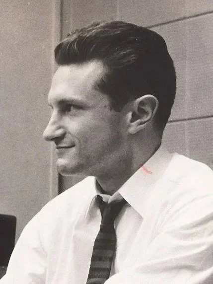 Black-and-white photo of a young man with slicked-back hair wearing a dress shirt and tie, sitting indoors.