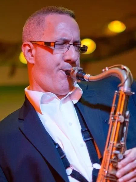 A man in a suit and glasses playing a saxophone on stage under warm lighting.