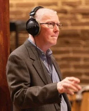 A man wearing glasses and headphones, standing in front of a brick wall.
