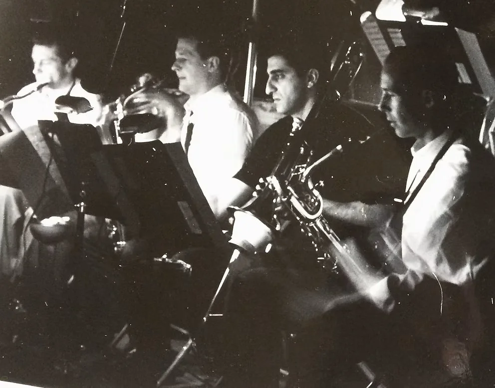 Four musicians playing involving saxophones, seated in a row with music stands and microphones, in a darkened setting.