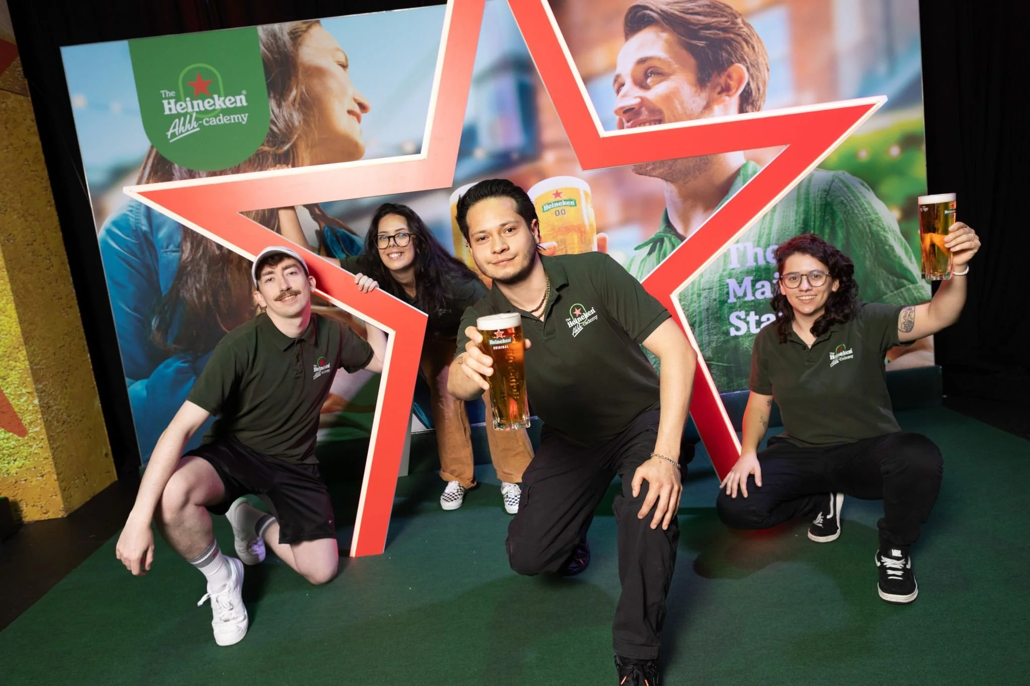 Four people posing with glasses of beer at a Heineken event, behind a colorful backdrop featuring large images of smiling people and the Heineken logo.