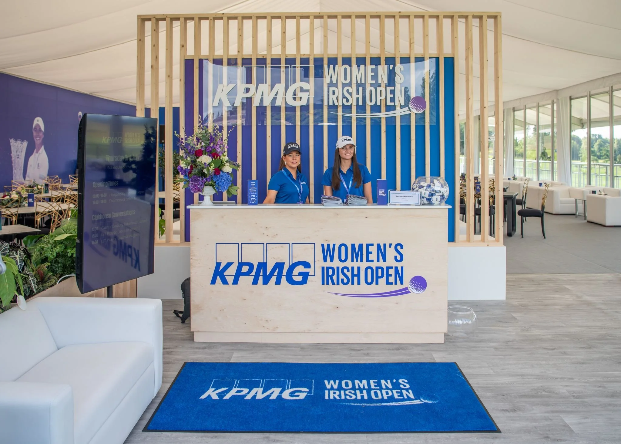 Reception desk with two women in blue shirts and caps at the KPMG Women's Irish Open event, with a blue banner behind them displaying the event name and logo, inside a well-lit indoor venue.
