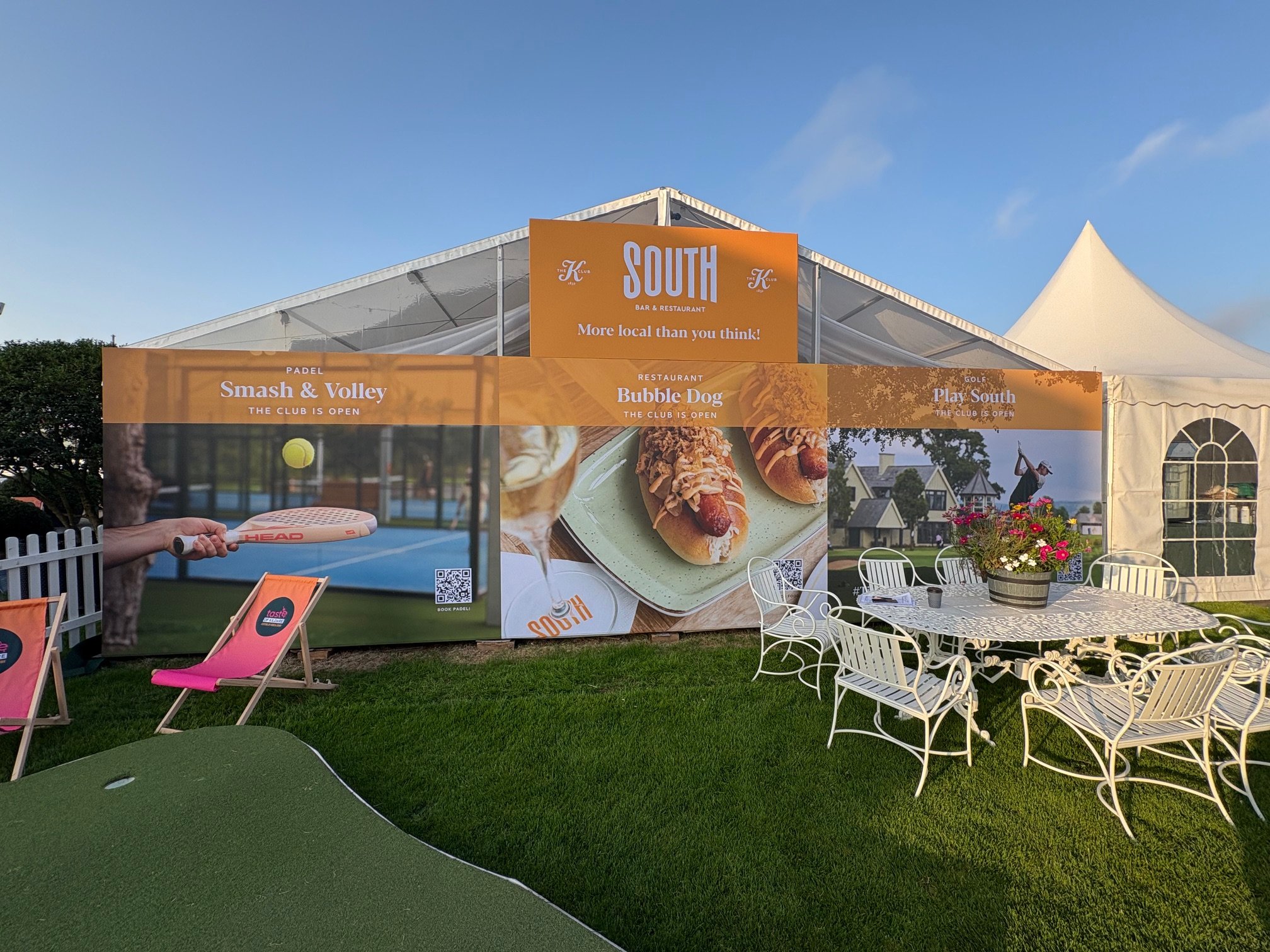An outdoor event setup with a white tent, a large orange banner displaying various activities including pickleball, hot dogs, and golf, surrounded by white chairs and a circular table with a flower arrangement. The sky is clear and the grass is green