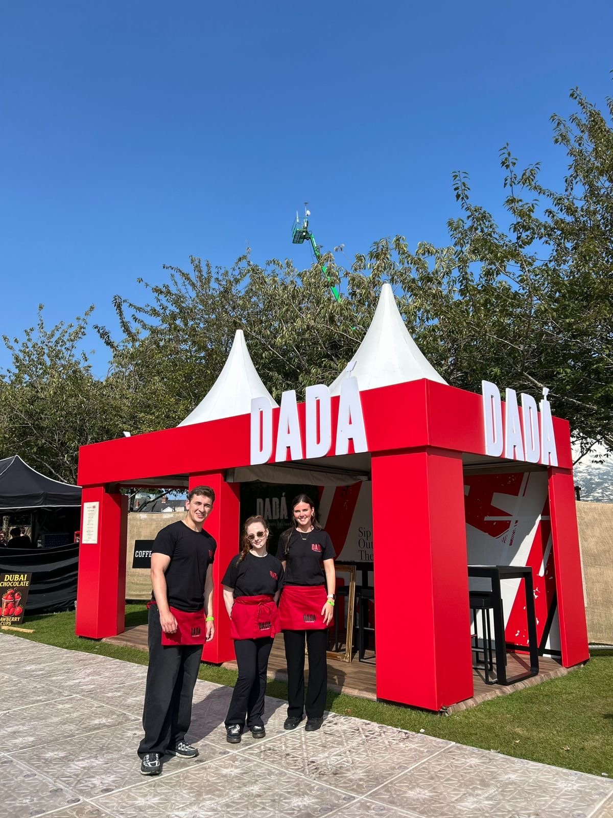 Three people standing in front of a red DADA tent at an outdoor event. They are wearing black shirts and red aprons. The sky is clear and blue, with some trees and a lift in the background.