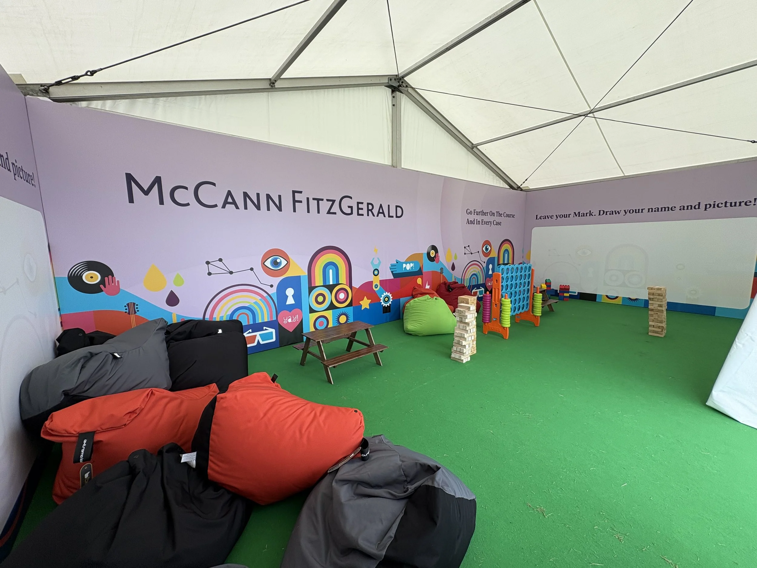 Children's play area with bean bags, colorful toys, and a green carpet, inside a tent with promotional branding for McCann FitzGerald.