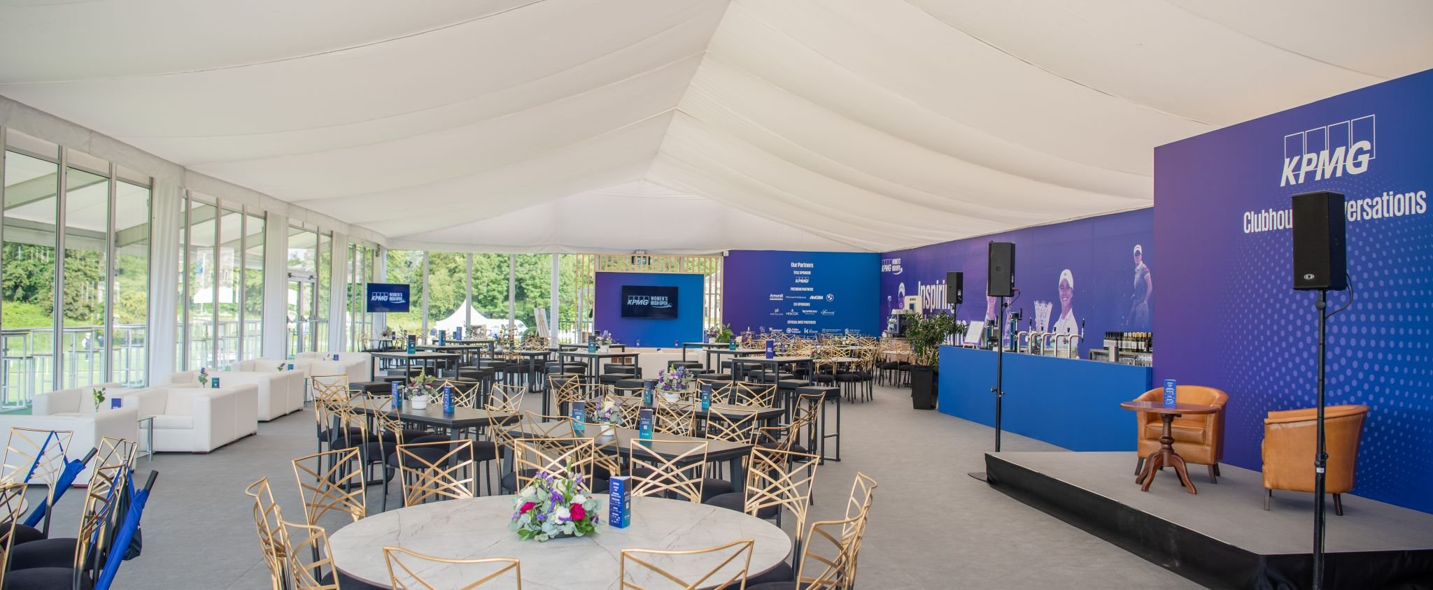 Interior view of a large event space set up for a conference with round tables, chairs, floral centerpieces, a stage with a large blue backdrop, and multiple screens displaying KPMG branding.