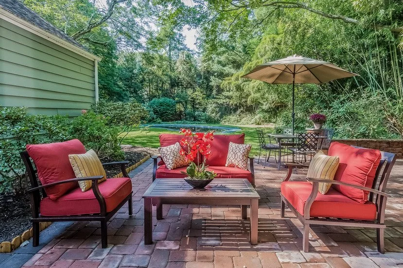 Outdoor patio area with red cushioned seating, a wooden table with a flower arrangement, a dining set with an umbrella, and lush green trees and bushes in the background.