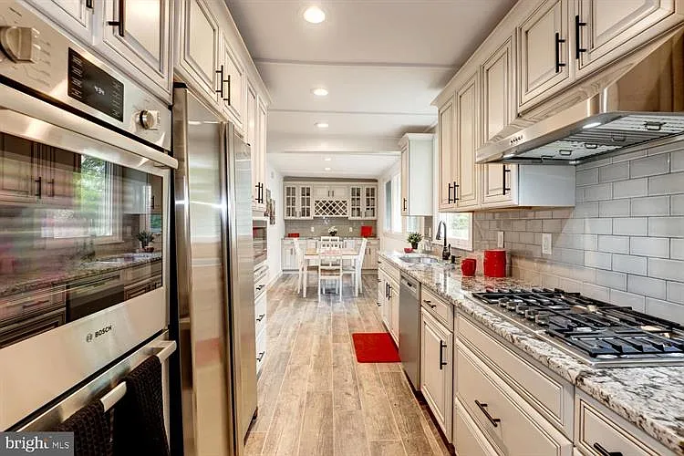 View of a modern kitchen with white cabinets, granite countertops, stainless steel appliances, and a dining area in the background. The kitchen has light wood flooring and red accents, including small rugs and decorative items.