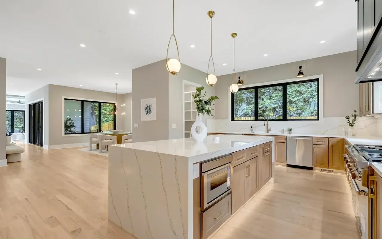 Modern kitchen with light wood cabinets, white marble countertops, and a large island with a vase of greenery. Black-framed windows offer a view of trees outside, and it's well-lit with pendant lights.