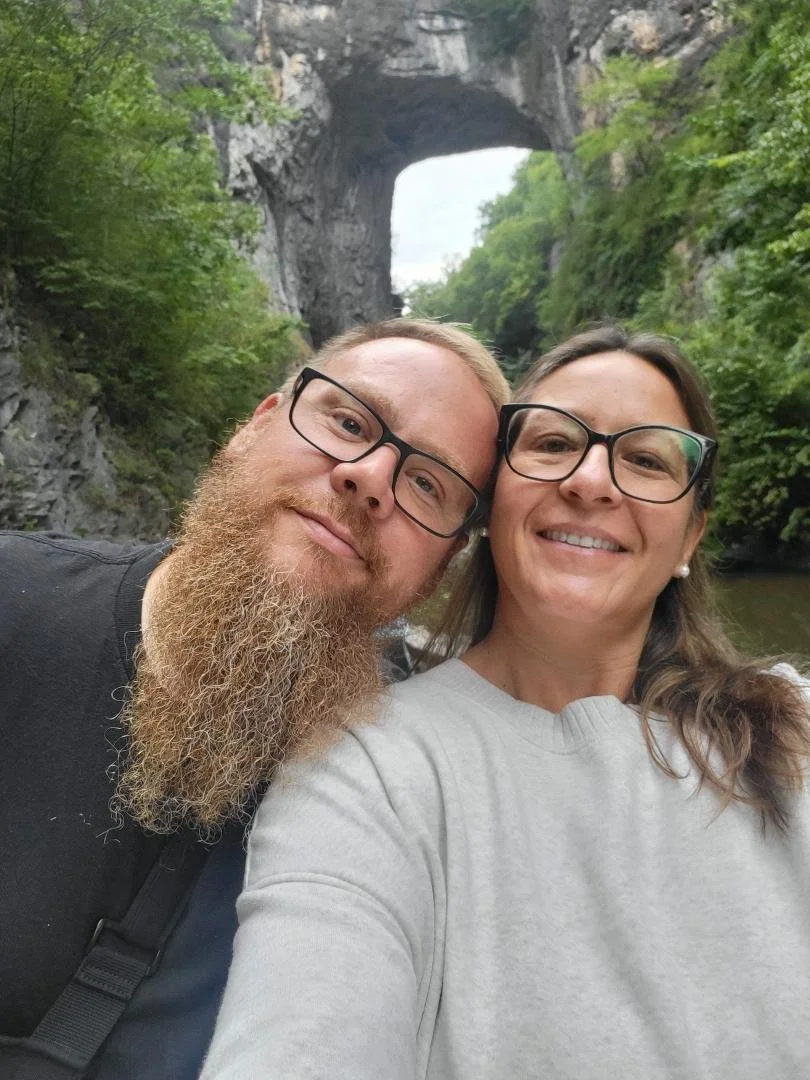 A couple taking a selfie outdoors near a river, with a large natural rock formation and an arch in the background, surrounded by green trees.