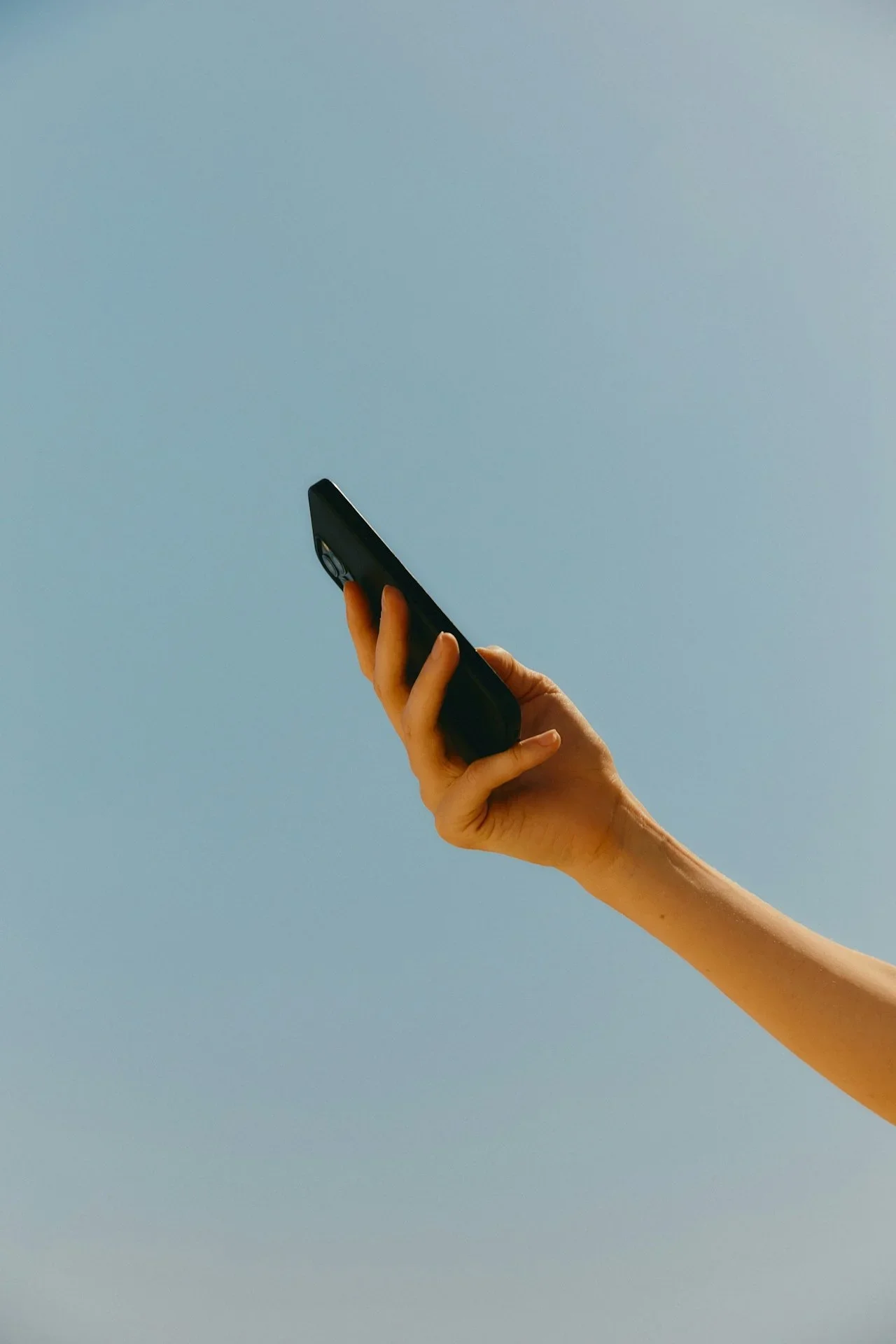 Close-up of a person holding a black mobile phone against a light blue sky.