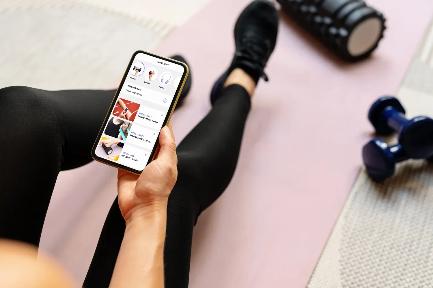 Person sitting on the floor in workout clothes, holding a smartphone that displays a fitness app, with dumbbells and a foam roller nearby.