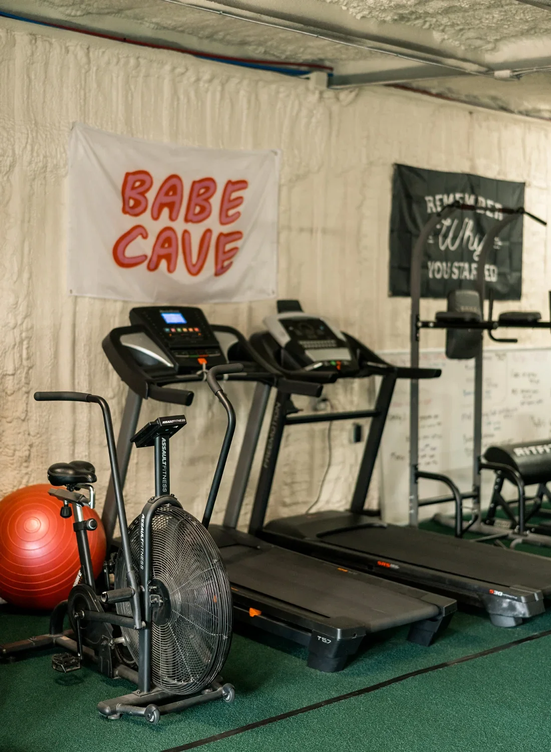 A small gym with two treadmills, an exercise bike, a red exercise ball, and workout banners on the wall.