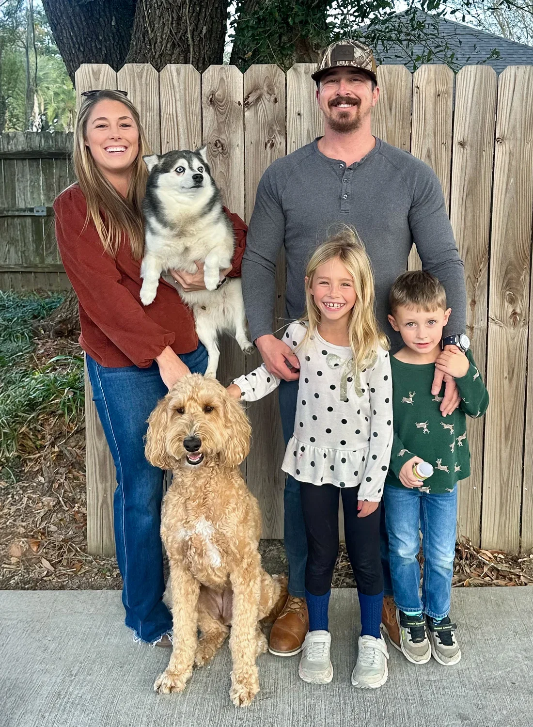 A happy family of four with two dogs, a dog being held by the woman, and another dog sitting on the ground, standing next to the girl. They are outdoors in front of a wooden fence and trees.