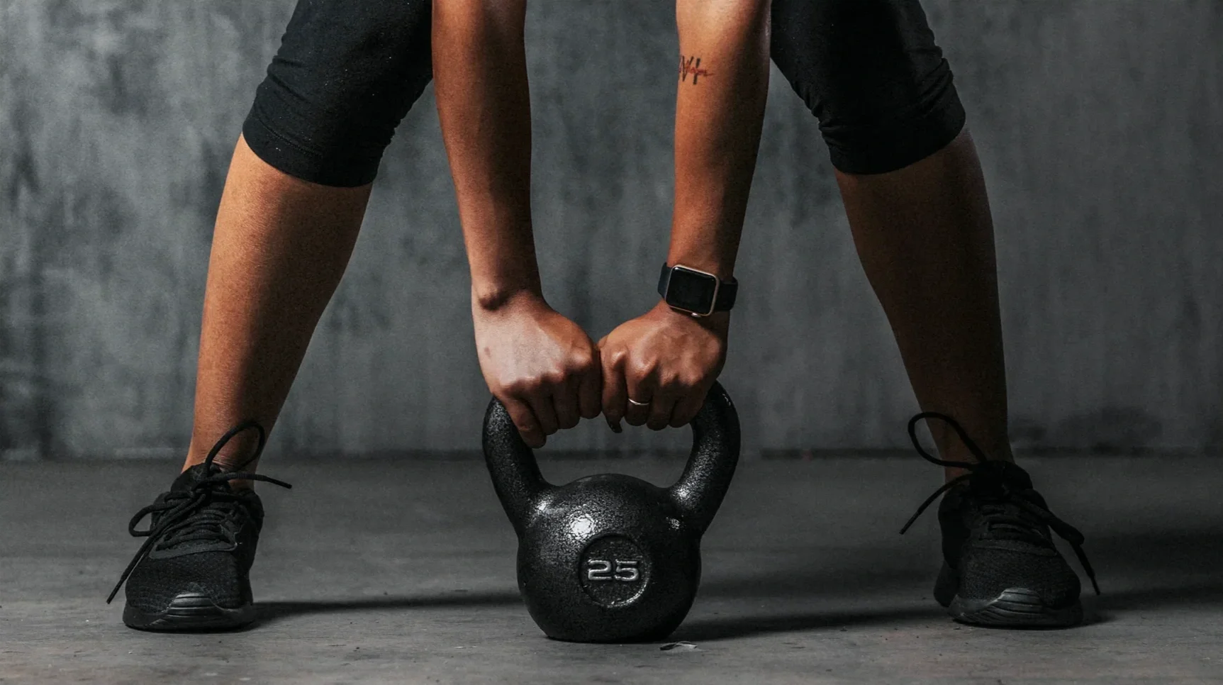 A person in black athletic clothing and shoes, wearing a smartwatch, is performing a workout with a black kettlebell, bent over with fists gripping the kettlebell handle in a gym setting.