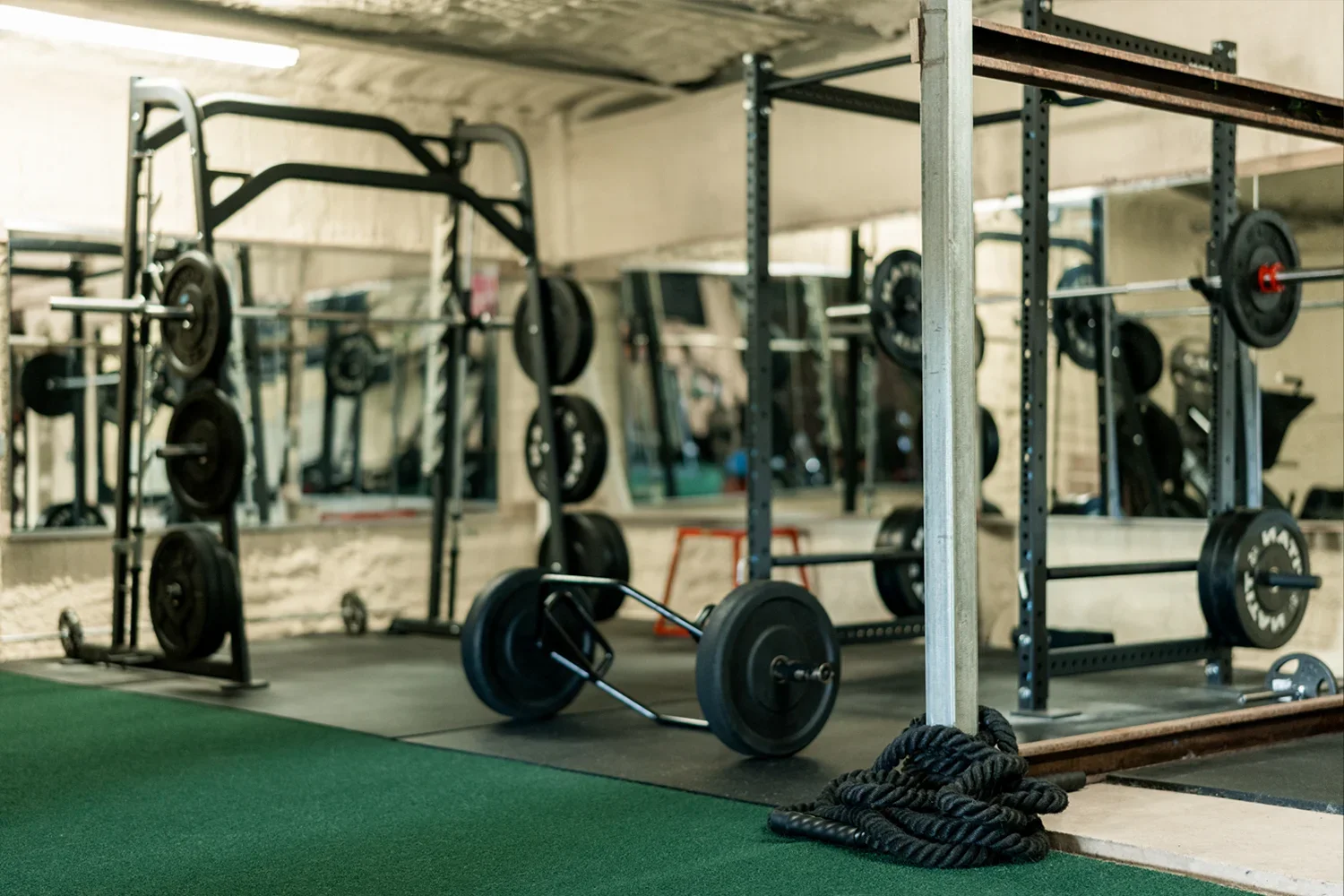 Workout gym with weightlifting equipment, including barbells, weight plates, squat racks, and a sledgehammer, on a black mat with a green turf area in the foreground.