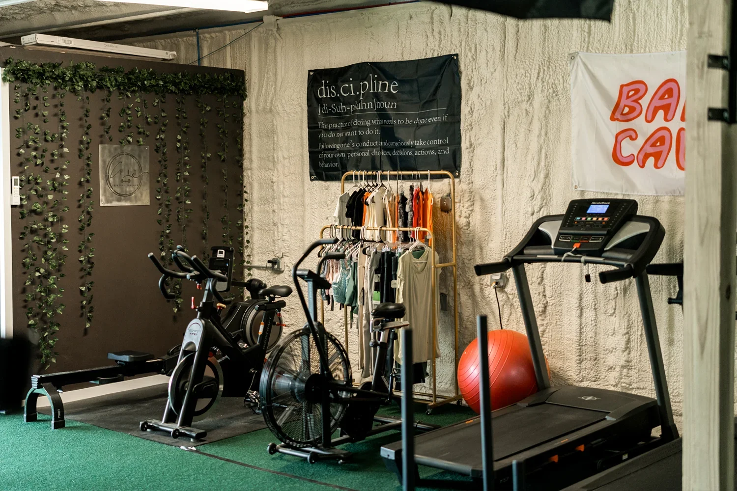 Indoor gym space with stationary bike, treadmill, orange exercise ball, and a rack of hanging clothes. Green carpet and wall decorations including a black sign with white text and green vines on a brown background.