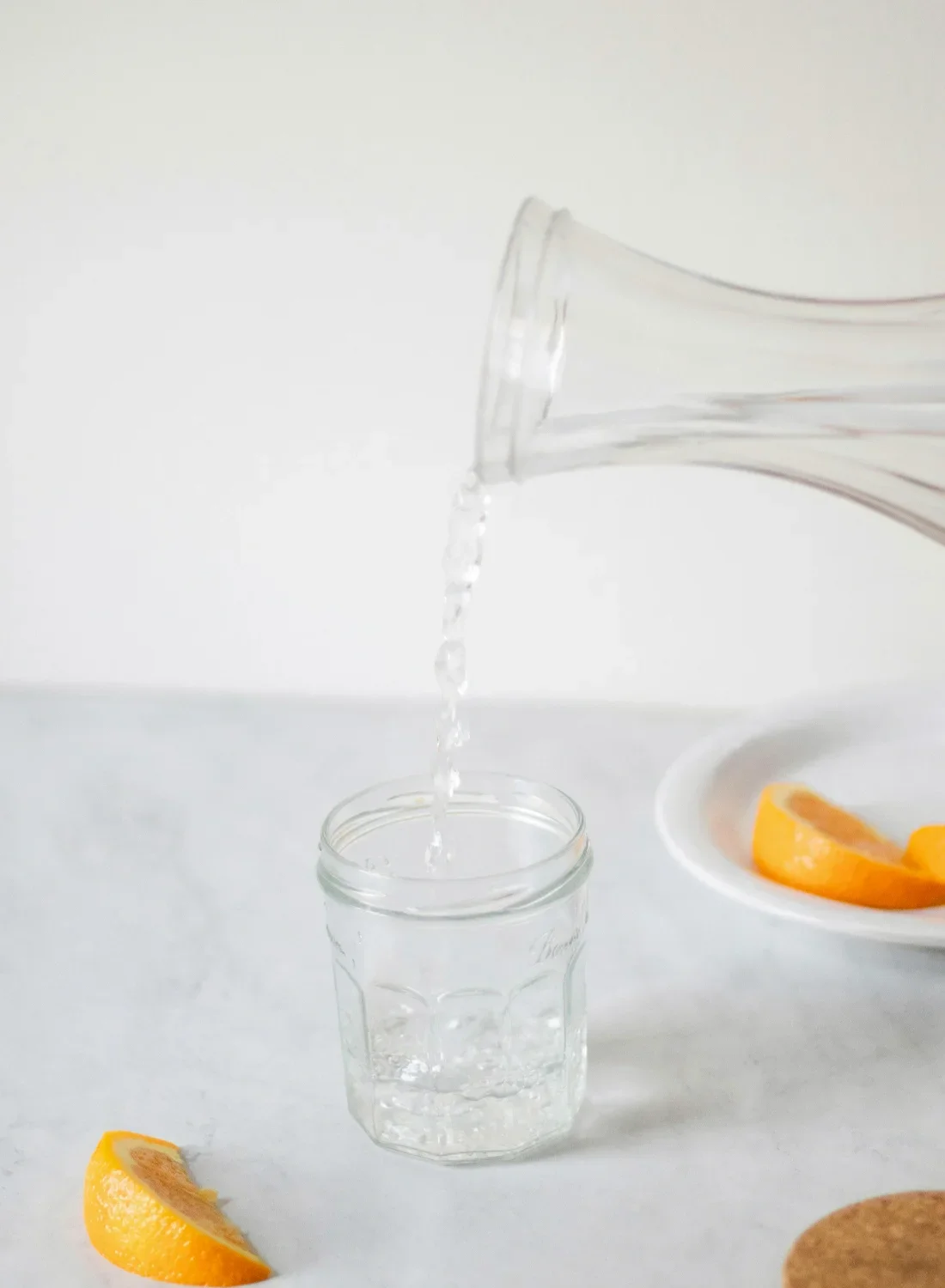 Water being poured from a glass pitcher into a clear glass jar on a white surface with orange slices on a white plate and nearby.