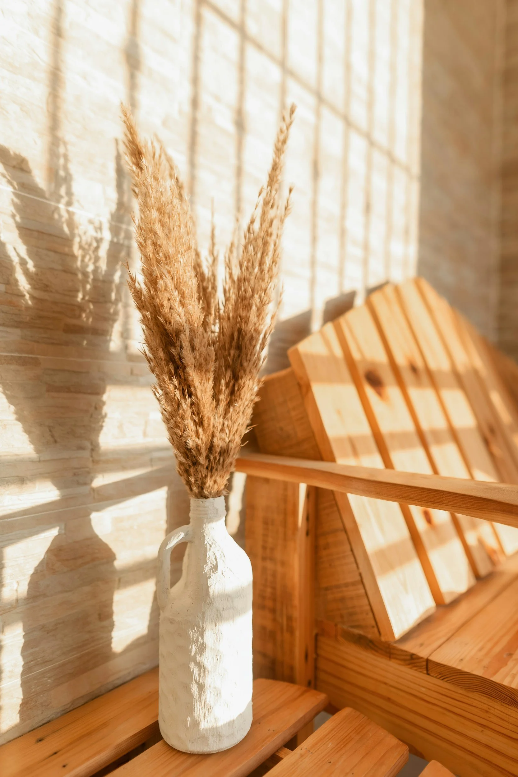 A white ceramic vase holding dried pampas grass on a wooden surface, with a stone wall and wooden slatted furniture in the background.