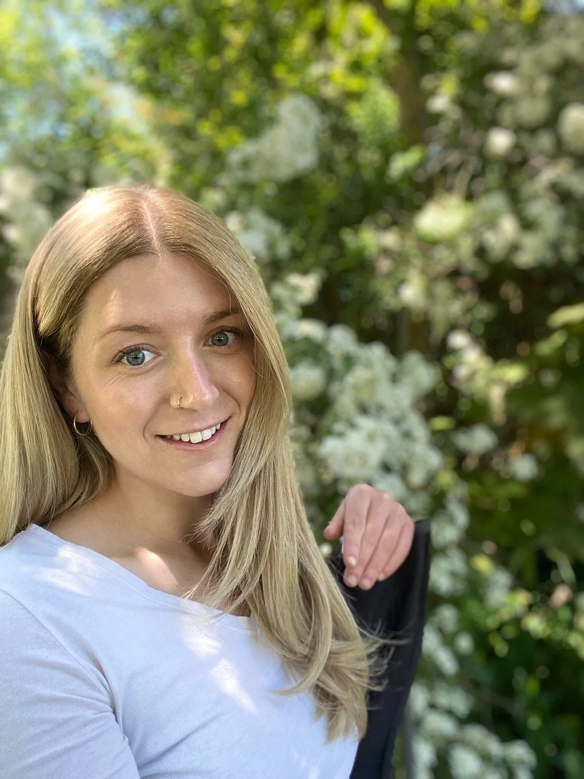 A young woman with blonde hair, blue eyes, and a nose ring smiling outdoors near white flowering bushes and green trees.