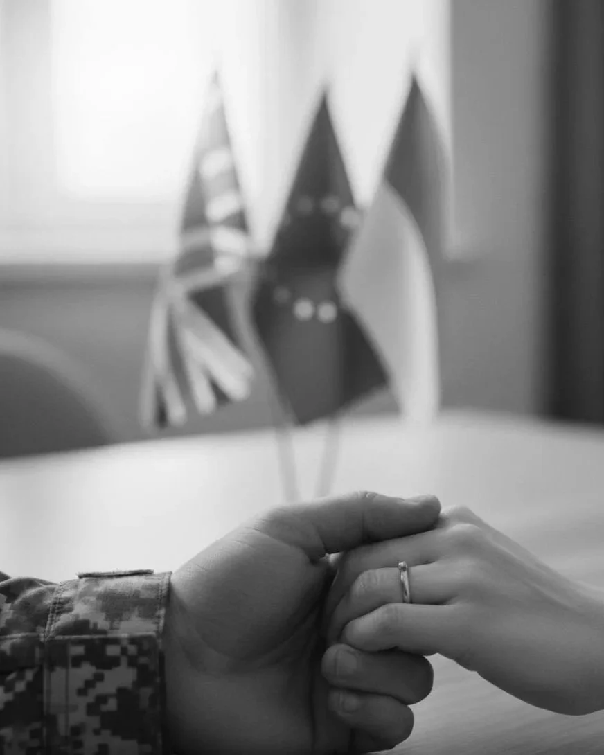 Two hands holding each other, with one wearing a ring, in front of three small flags including the American flag.