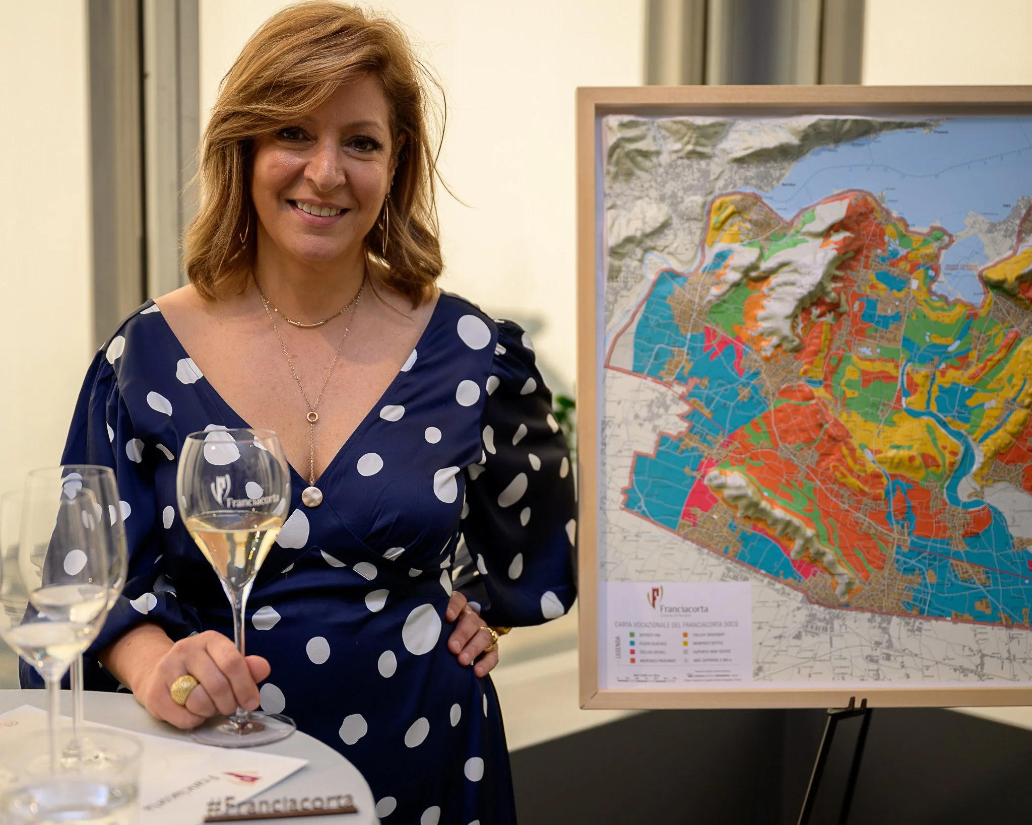 A woman with shoulder-length light brown hair smiling and standing next to a colorful map of a city or region on an easel, holding a glass of white wine, wearing a dark blue dress with white polka dots, and standing at a table with other glasses and 