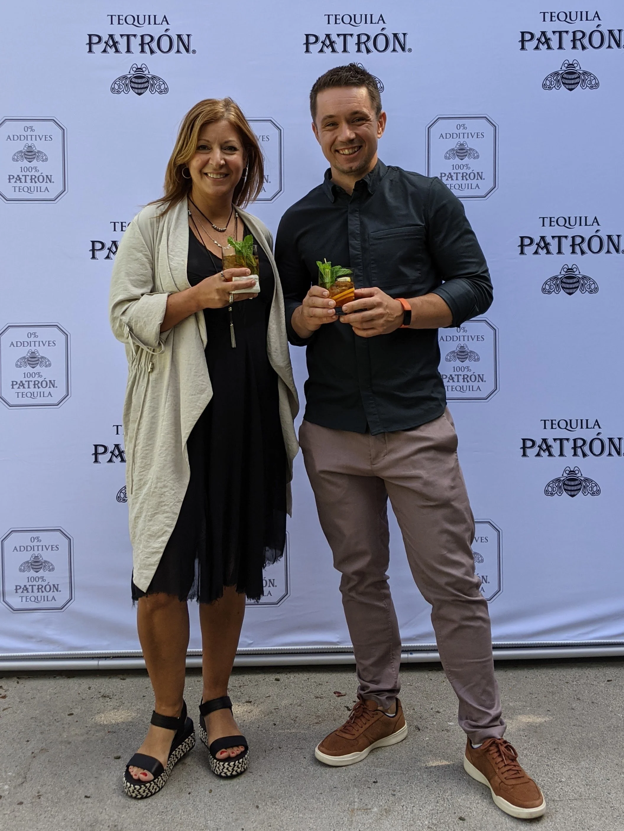 Two people holding drinks and smiling at an event with a Patrón tequila backdrop.
